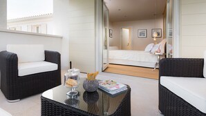 Un balcón con mobiliario de mimbre, una mesa de centro de vidrio con revistas, y una vista parcial de un dormitorio con cama y cuadros.