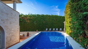 Una piscina rectangular con borde de azulejos, rodeada por un muro de piedra y un seto.