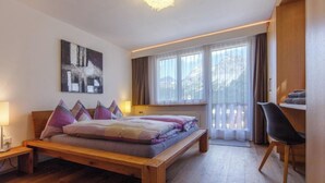 Une chambre à coucher avec un lit en bois, un bureau et une chaise. Il y a une fenêtre avec des rideaux et une vue sur l’extérieur.