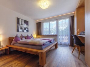 Ein Schlafzimmer mit einem hölzernen Bett, einem Schreibtisch und einem Stuhl. Ein Fenster mit Vorhängen bietet Blick ins Freie.
