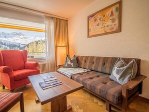 Una habitació acollidora amb un butac armchair vermell, una taula de cafè de fusta amb revistes, un sofà estampat amb coixins, vista a les muntanyes per la finestra, i un quadre a la paret.