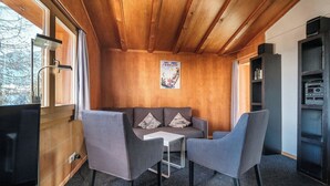 Ein gemütliches Zimmer mit Holzbalkendecke, einer Couch, zwei Stühlen, einem kleinen Tisch und einem Bücherregal.