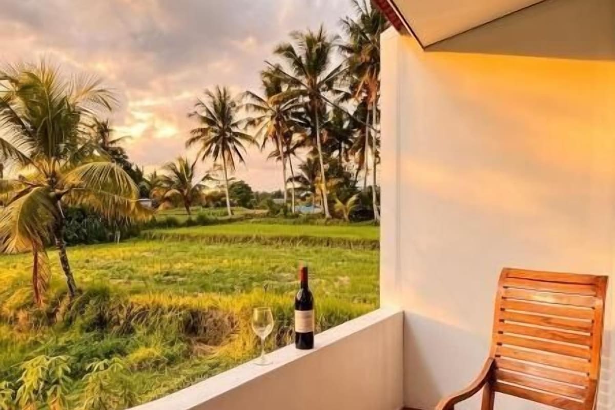 Un balcón con una silla de madera, una botella de vino y vistas a un campo verde y palmeras.