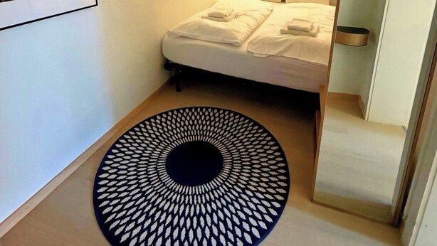 Ein Schlafzimmer mit einem Bett, einem an der Wand montierten Fernseher, einem gerahmten Bild an der Wand und einem runden Teppich auf dem Boden.