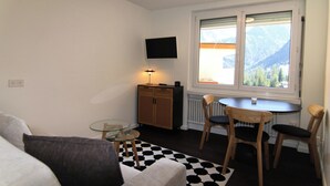 Ein kompakter Wohnraum mit einer Couch, einem kleinen runden Tisch, einem Schrank und einem Fenster mit Blick auf die Berge.