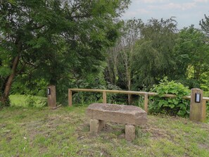 Eine Steinsitzbank in einem bewaldeten Gebiet, mit einem Holzzaun und zwei Hinweisschildern.