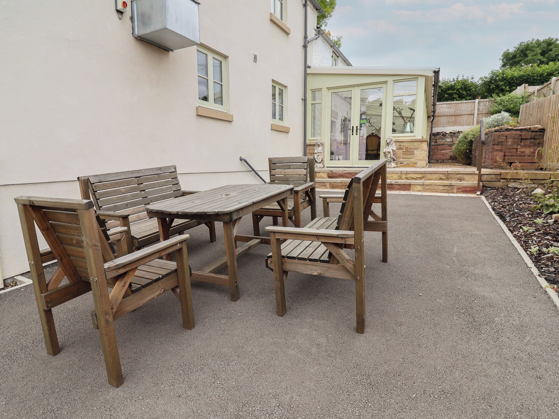 A patio with a wooden table and chairs, a white wall, and a glass door leading to a garden.