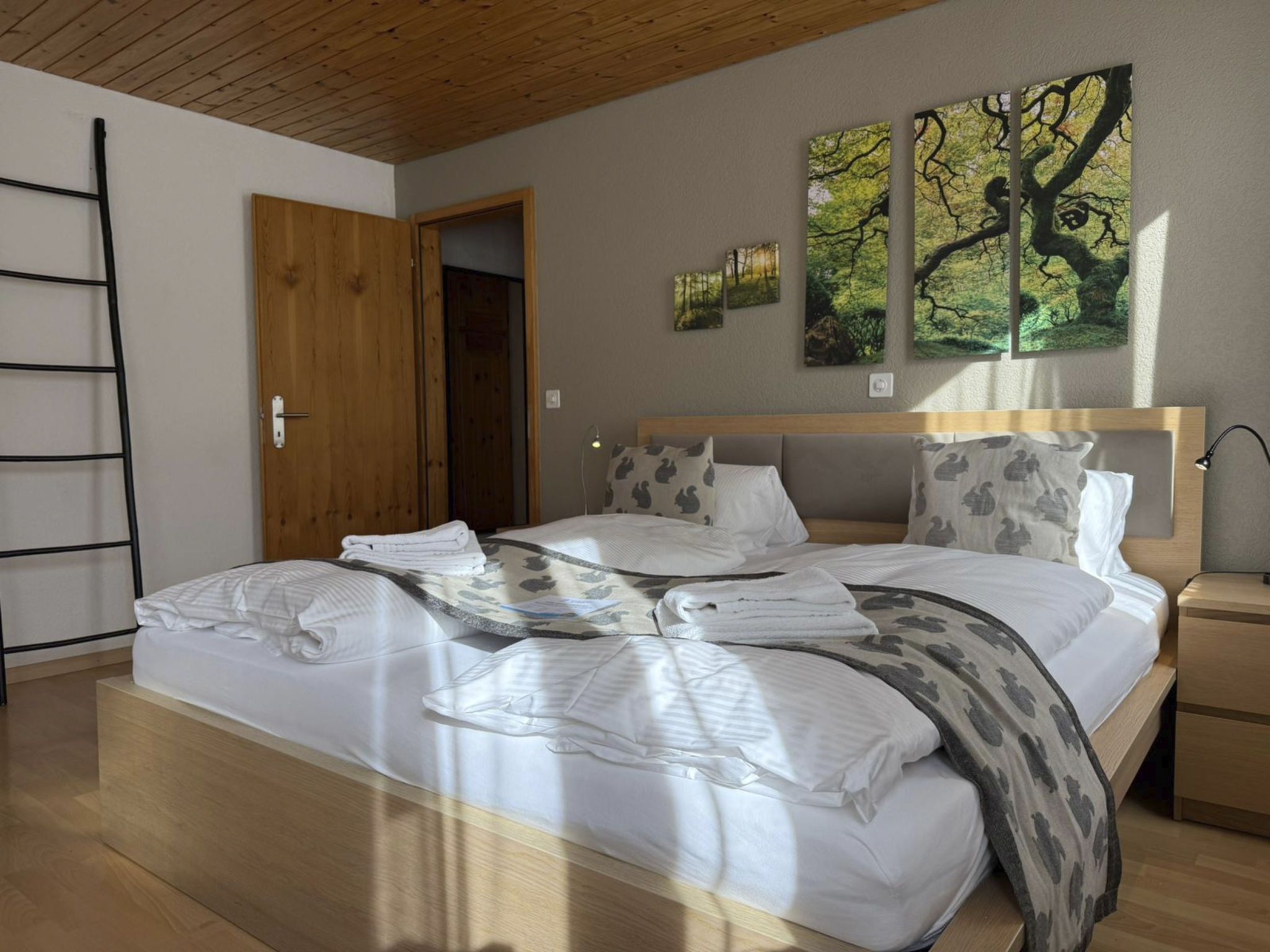 Una cama bien tendida con almohadas y una manta, una puerta de madera, una escalera y una pared con tres cuadros.