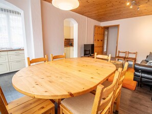 Ein Essbereich mit einem großen Holztisch, umgeben von Stühlen, einem Fernseher und einer Couch in einem Raum mit Holzbalkendecken und weißen Wänden.
