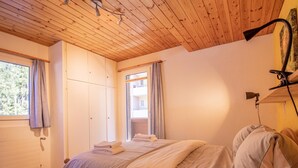 Une chambre avec un plafond en bois, un lit recouvert d’une couvre-lit à rayures, une fenêtre avec des rideaux et une vue sur un bâtiment à l’extérieur.