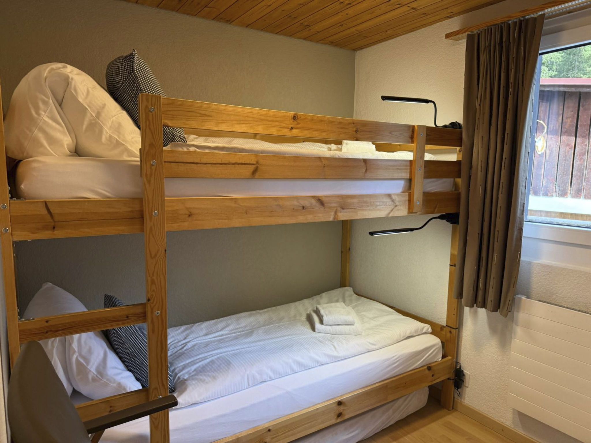 Uma cama beliche com dois níveis, uma janela com cortinas e teto de madeira.