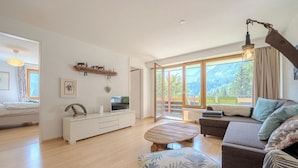 Ein geräumiges Wohnzimmer mit einer Couch, einem Couchtisch und einem Fernseher auf einem weißen Schrank. Große Fenster bieten einen Blick ins Freie.