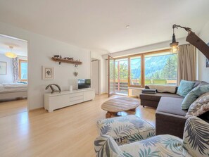 A spacious living room with a sofa, a coffee table, and a TV on a white cabinet. Large windows offer a view of the outdoors. - Marlenca 2 by Arosa Vacations Appartement/Fewo, Dusche und Bad, WC, 2 Schlafräum by Interhome (Arosa)