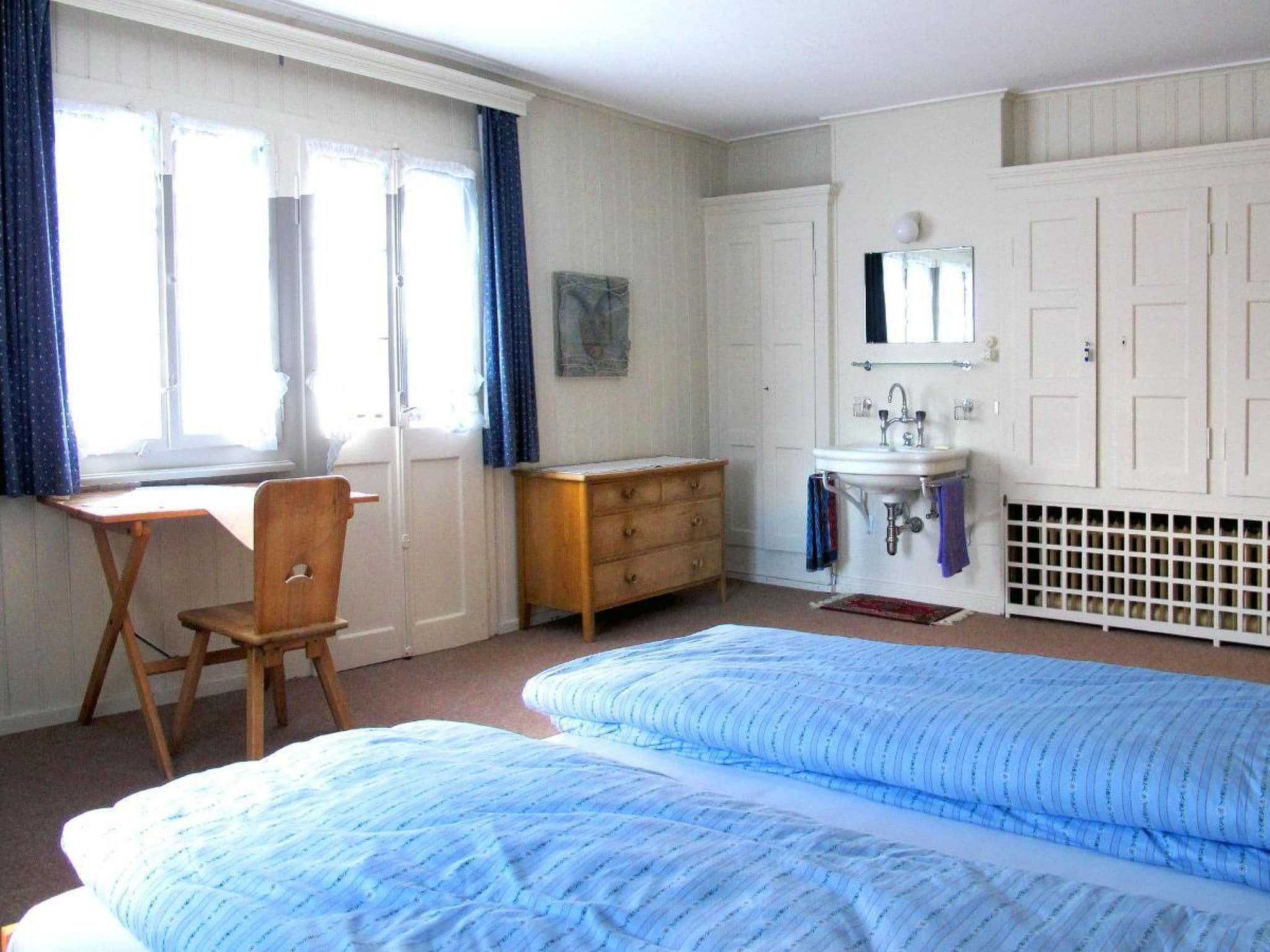 A bedroom with two beds, a dresser, a desk, a window with curtains, and a bathroom with a sink and mirror.