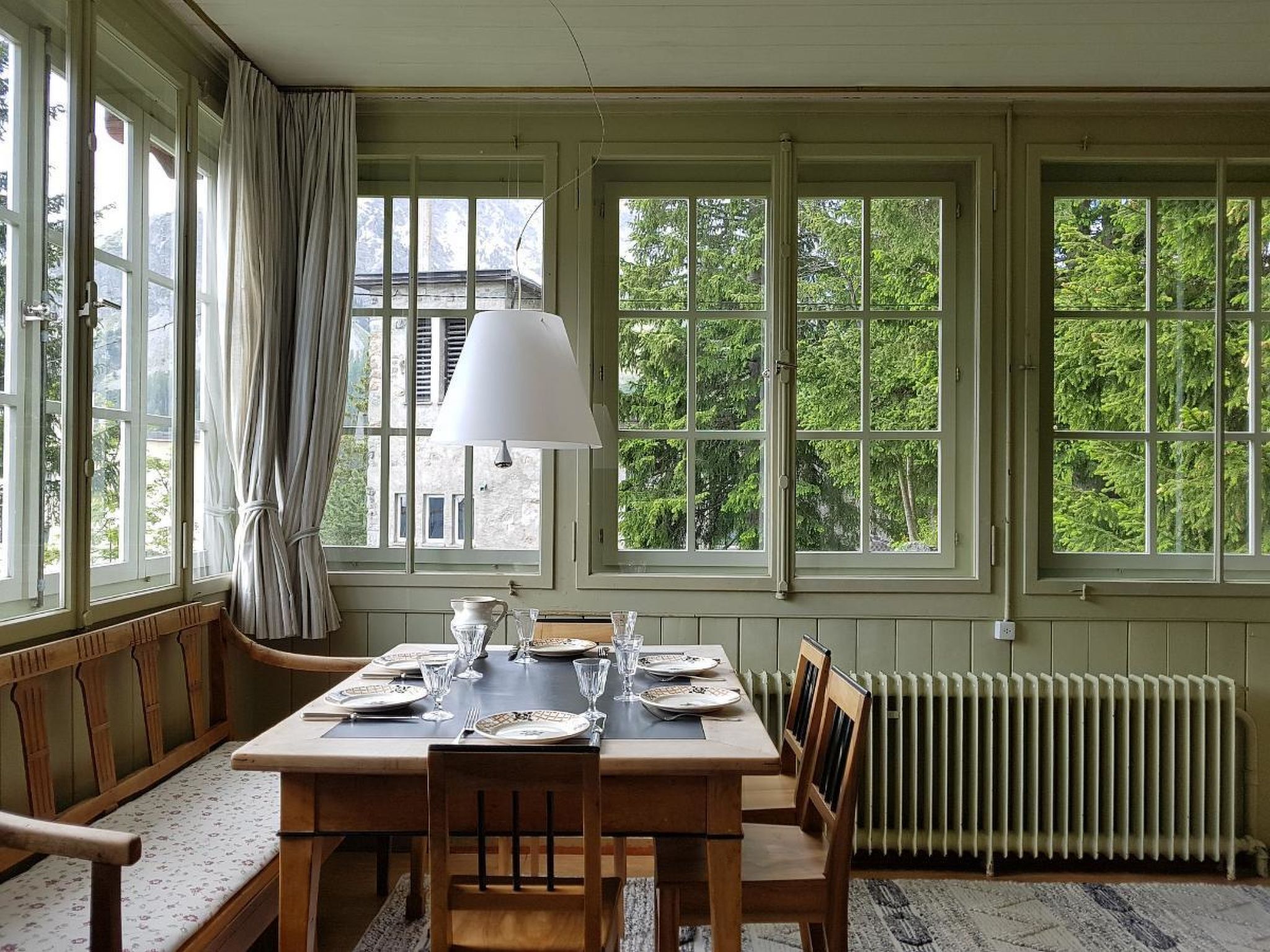 Comedor con una mesa lista para cuatro personas, ventanas amplias y un radiador.