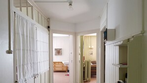 Ein schmaler Flur mit einem Bett im Hintergrund, einem Heizkörper und einem Regal mit Gegenständen auf der rechten Seite.