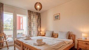 Ein Schlafzimmer mit einem hölzernen Bett, einem Nachttisch, einem Sessel, einem Fenster mit Vorhängen und einem Bild an der Wand.