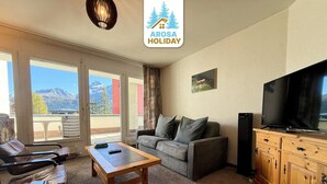 Ein Wohnzimmer mit einer Couch, einem Holztisch, einem Fernseher und Bergblick durch große Fenster.