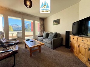 Ein Wohnzimmer mit einer Couch, einem Holztisch, einem Fernseher und Bergblick durch große Fenster.