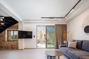 A modern living room with a sofa, a coffee table, a flat-screen TV mounted on the wall, and a staircase leading to an upper level. - Marvelous new Luxury Villa With Heated Pool! (Rethymno)