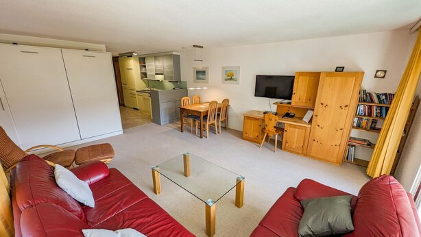 Un espacio habitable compacto con un sofá rojo, una mesa de centro de vidrio, zona de comedor y cocina con armarios de madera.
