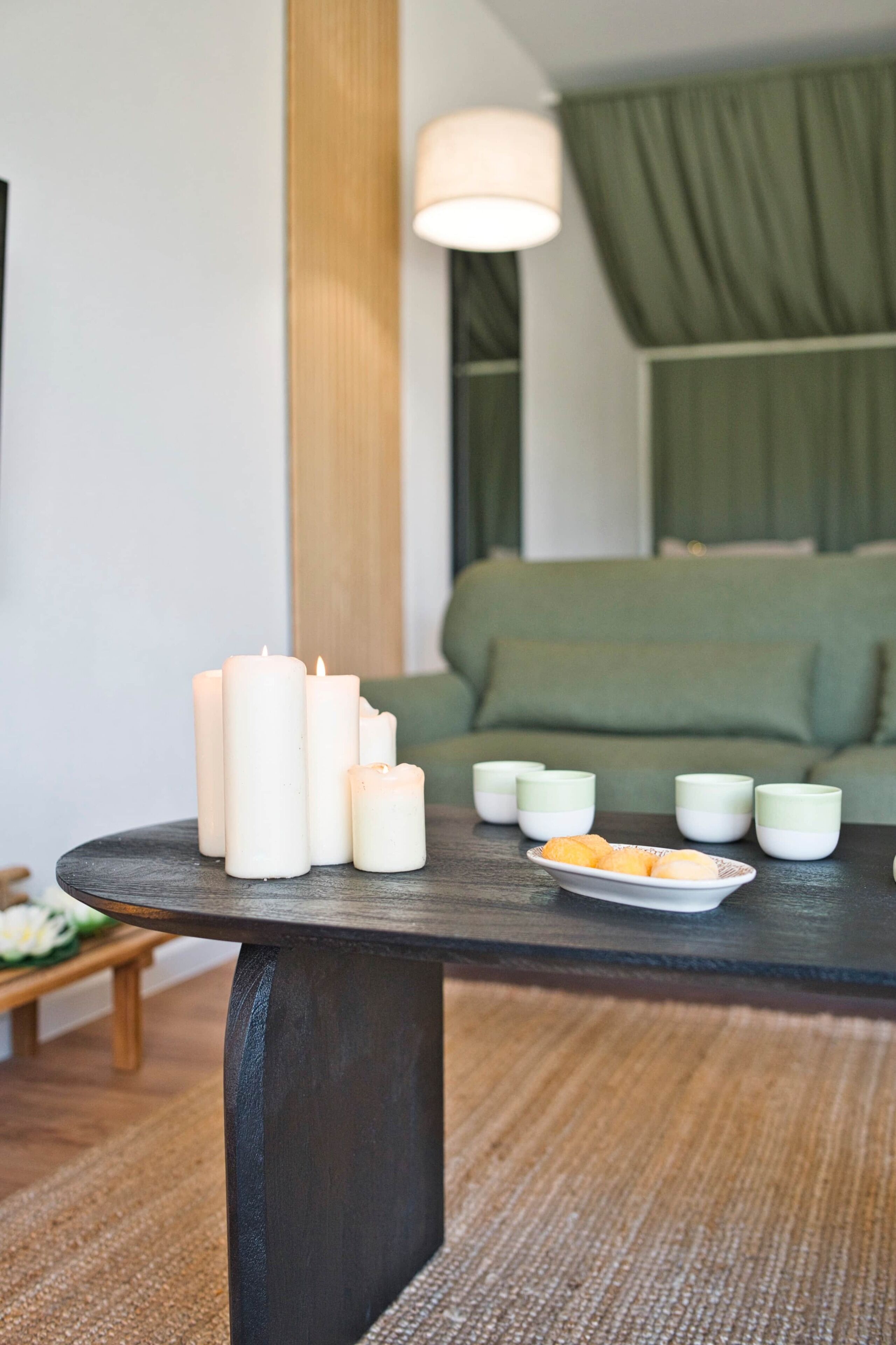 Una habitación con una mesa de madera oscura que sostiene velas y tazas, un sofá verde y una pared blanca.