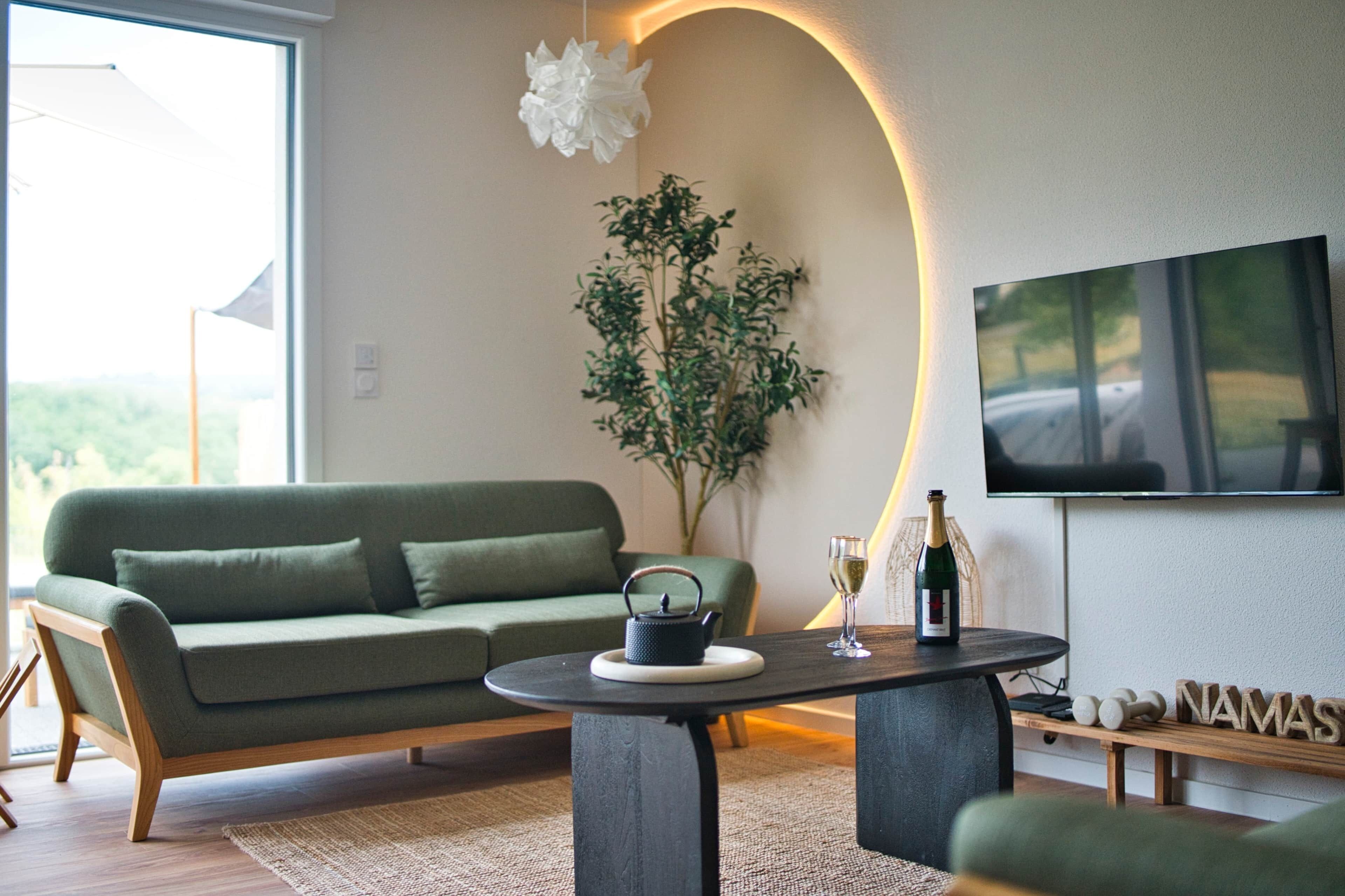 A modern living room with a green sofa, a coffee table, a potted plant, a bottle of champagne, and a flat-screen TV mounted on the wall.