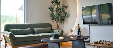 A modern living room with a green sofa, a coffee table, a potted plant, a bottle of champagne, and a flat-screen TV mounted on the wall.