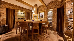A brick-walled room with a wooden table and chairs, a hanging light fixture, and shelves with bottles.