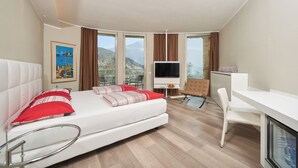 A modern hotel room with a bed, TV, desk, and large windows offering a mountain view.