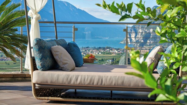 A balcony with a cushioned seating area, a wicker chair, and a view of mountains and a town.