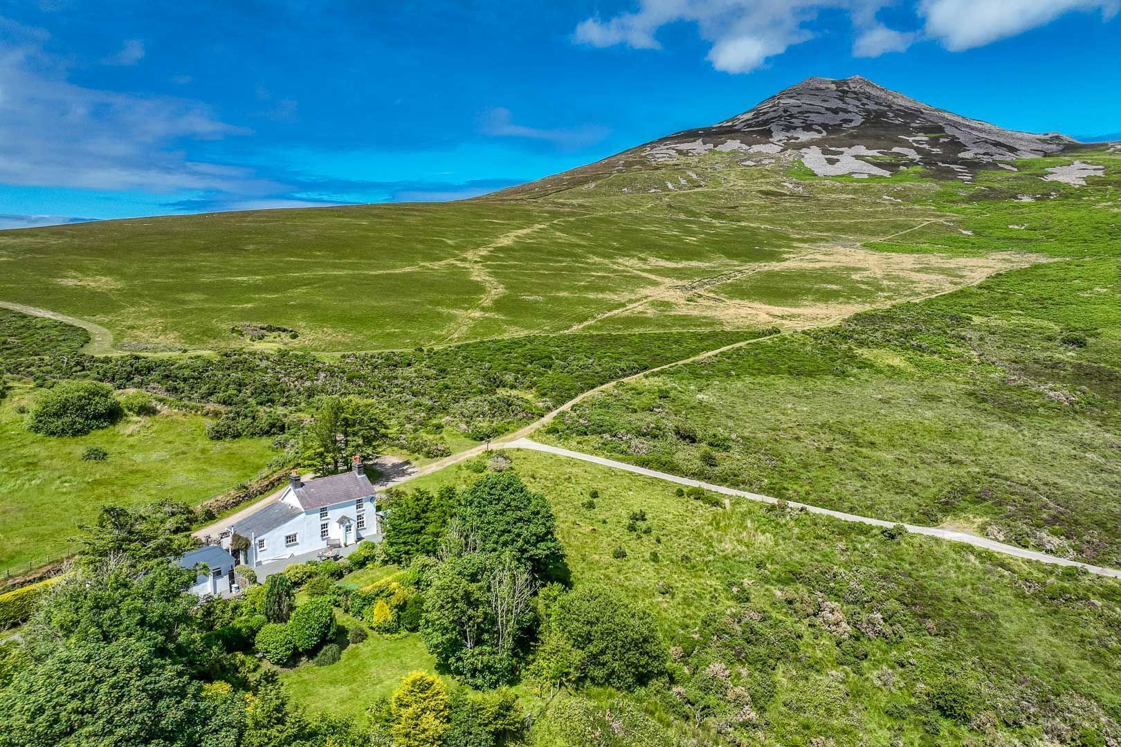 Une maison avec une allée pavée, entourée de champs verdoyants et avec une montagne en toile de fond.