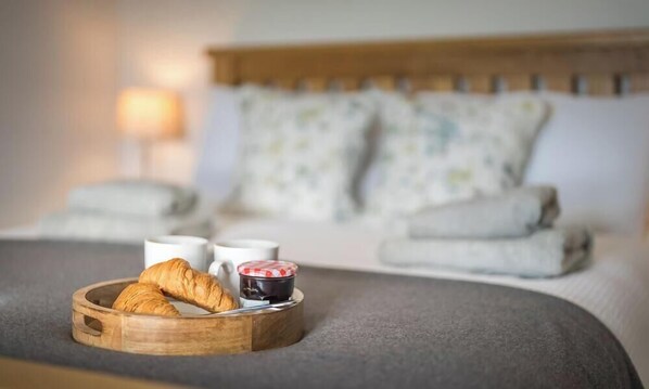 Una cama con una manta gris, una bandeja de madera con cruasanes y un tarro de mermelada, y una lámpara de noche.