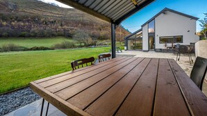 Mesa e cadeiras de madeira sob um alpendre, com vista para uma casa moderna e paisagem gramada.