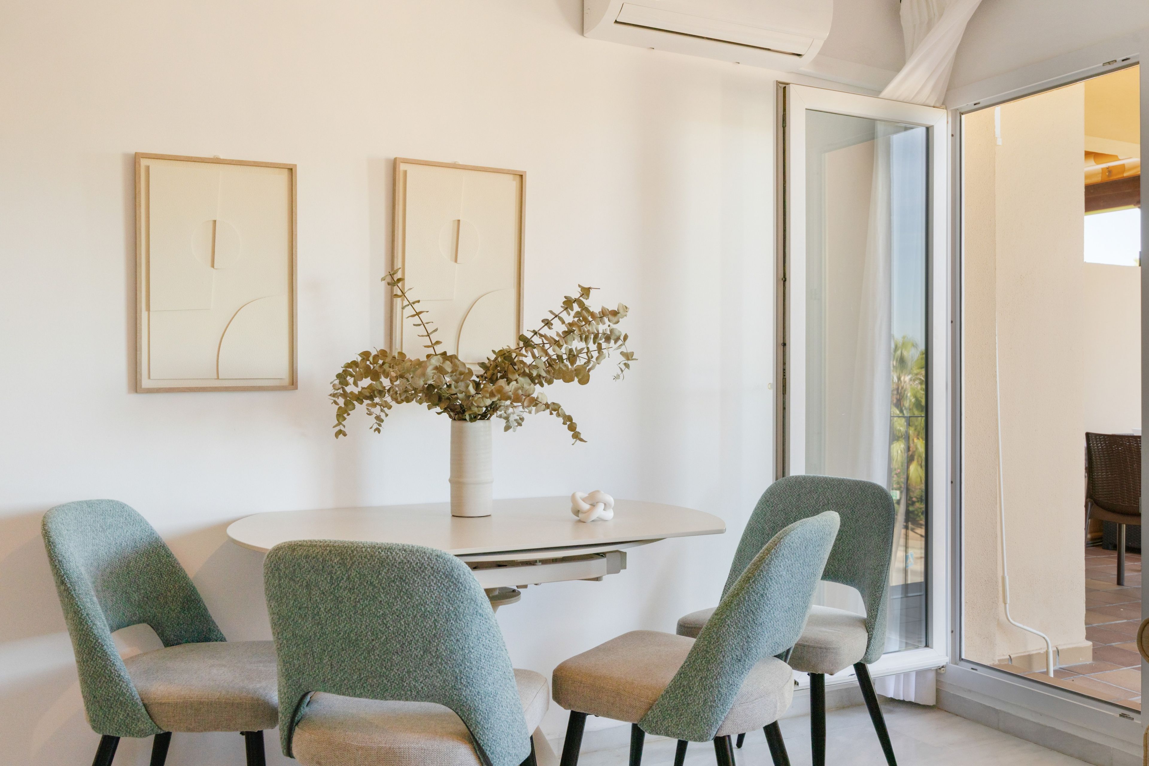 Uma área de refeições com uma mesa redonda, quatro cadeiras, duas obras de arte emolduradas e um vaso com flores secas.