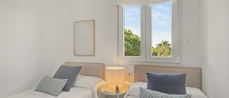 A bedroom with two beds, a bedside table with a lamp, a window with a view of greenery, and a framed picture on the wall.