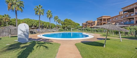 Una piscina con un sendero de ladrillo que la atraviesa, rodeada de palmeras y un edificio con balcones.