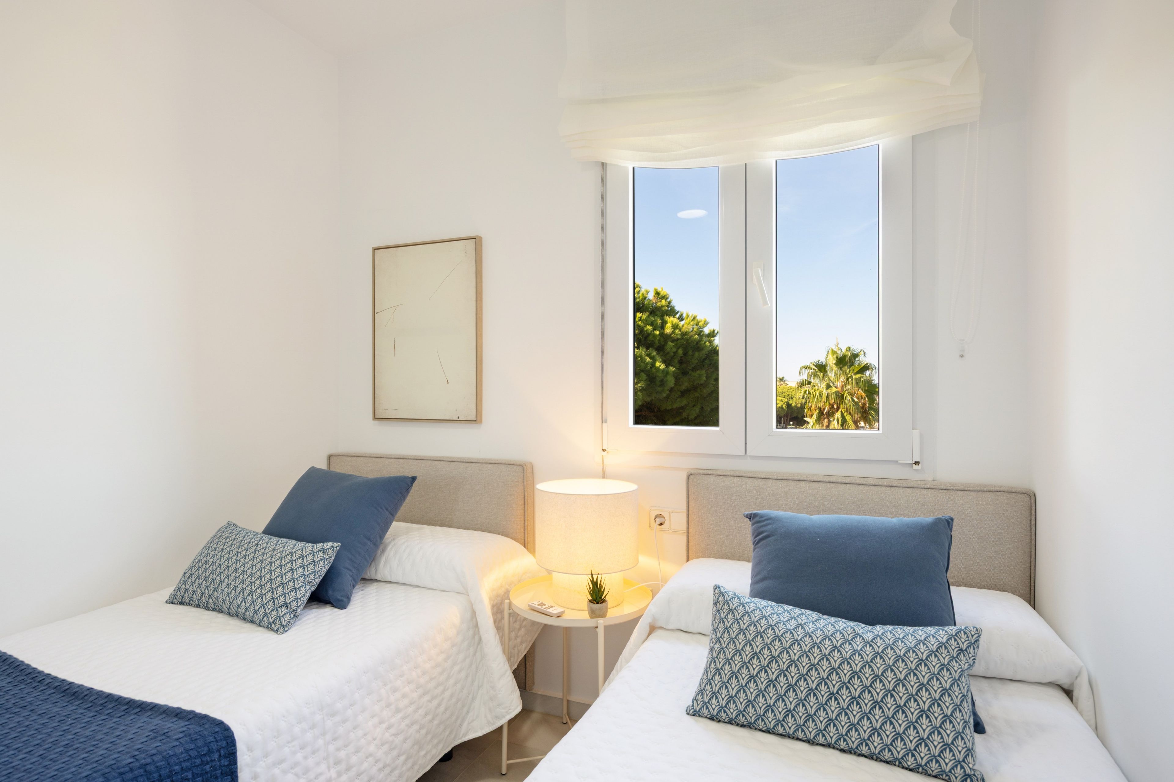 Quarto de hotel com duas camas, mesa de cabeceira com abajur, janela com vista para o verde e quadro na parede.