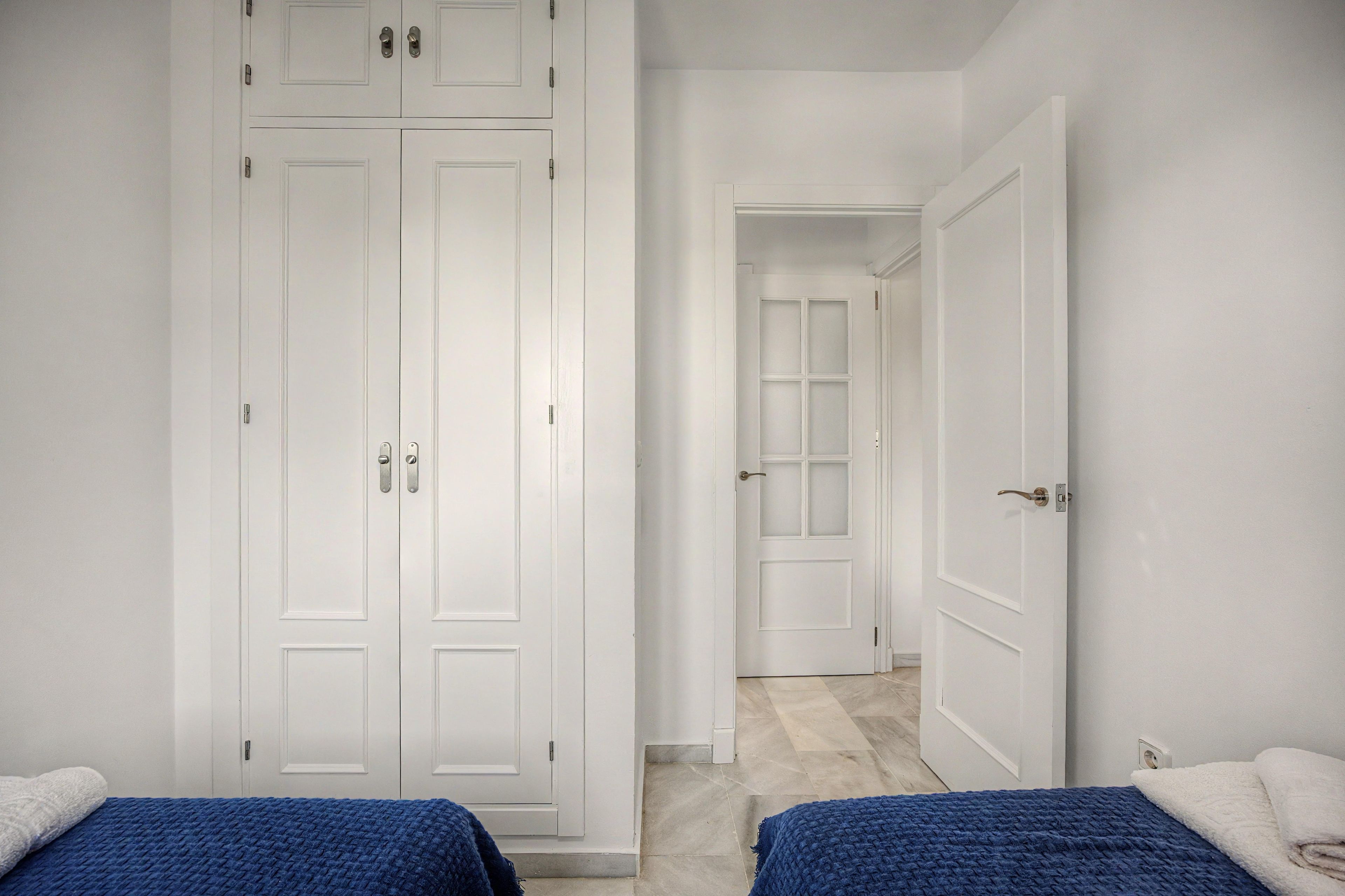A room with a large wardrobe, a bed with a blue blanket, and a white door leading to another room.