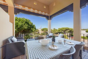 Un área de comedor al aire libre techada, con manteles a cuadros, mobiliario de mimbre y vistas de la vegetación y edificios circundantes.