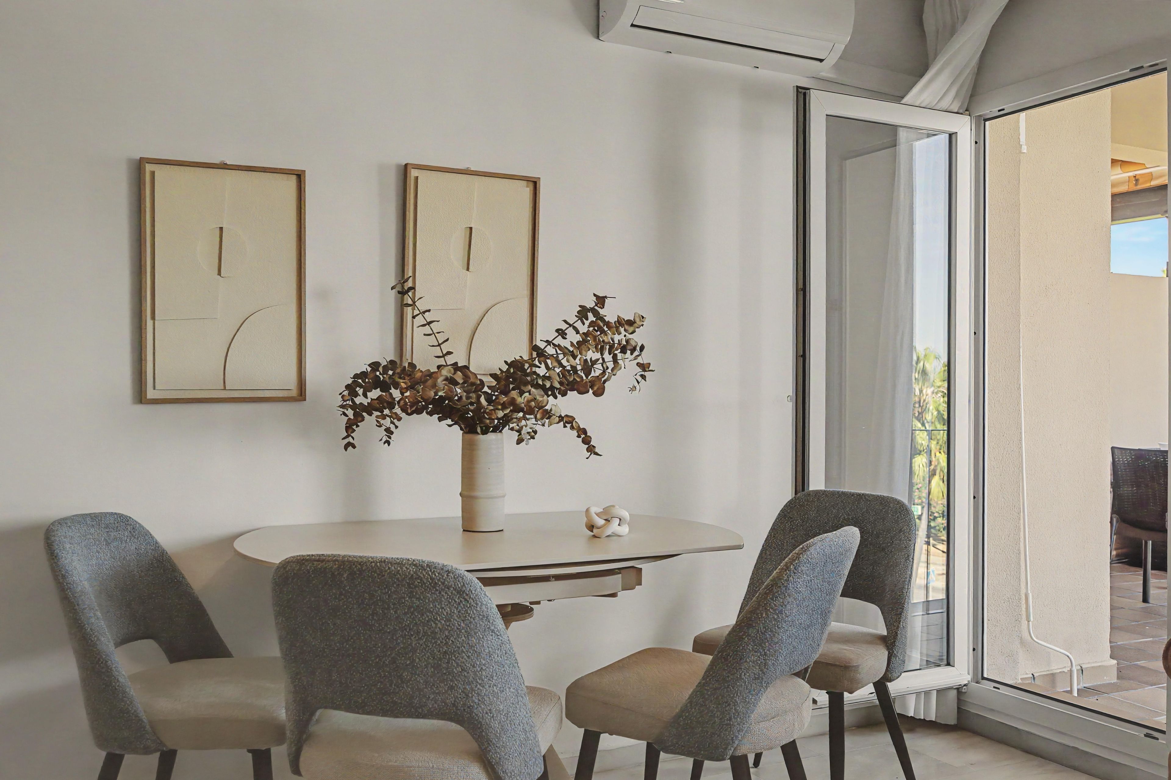 A dining area with a round table, four chairs, two framed artworks, a vase with dried plants, and a sliding glass door leading to an outdoor space.