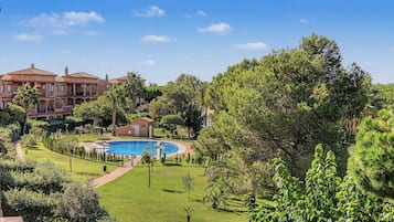 Una piscina rodeada por un césped cuidado, con un árbol grande al frente y un edificio al fondo.