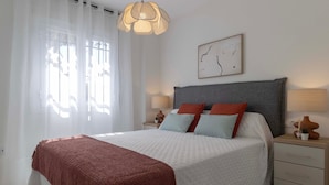 Un dormitorio con cama, lámparas de noche, mesita de noche, ventana con cortinas translúcidas y una obra de arte en la pared.