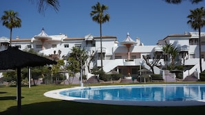 Una piscina rodeada de edificios blancos con balcones, palmeras y un paraguas de paja.