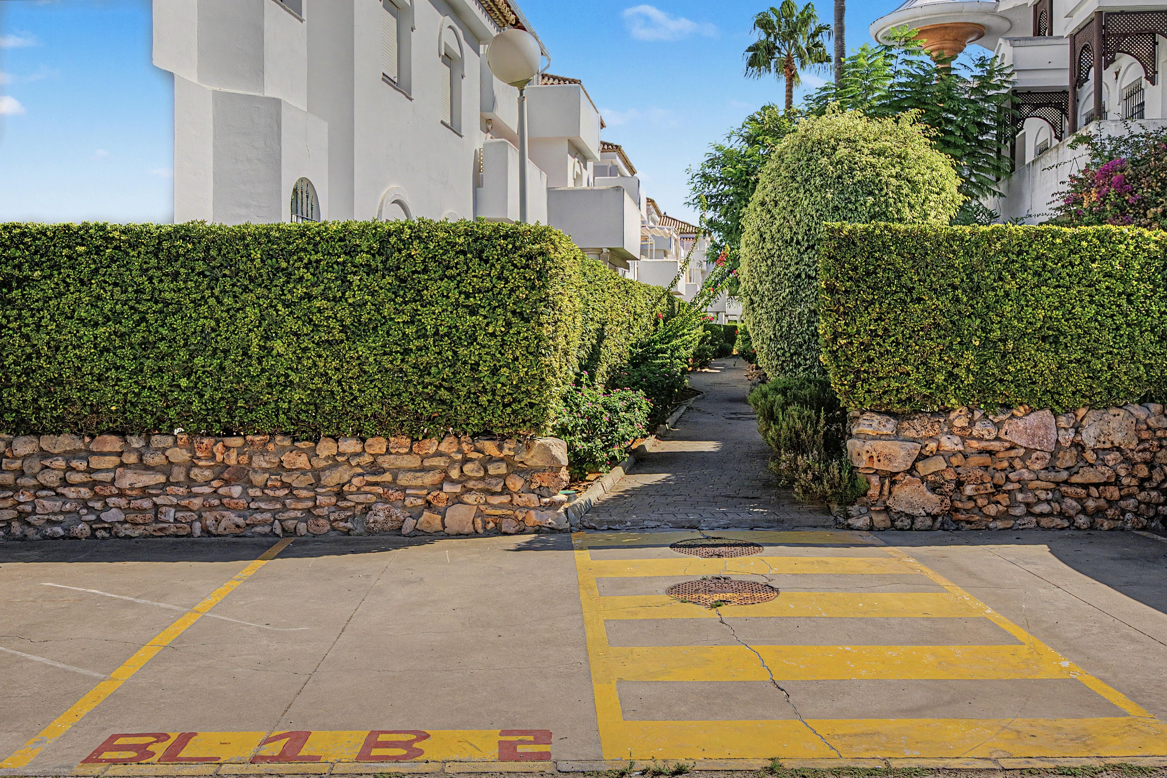 Una calle estrecha bordeada por muros de piedra y setos, que conduce a un edificio con balcones.