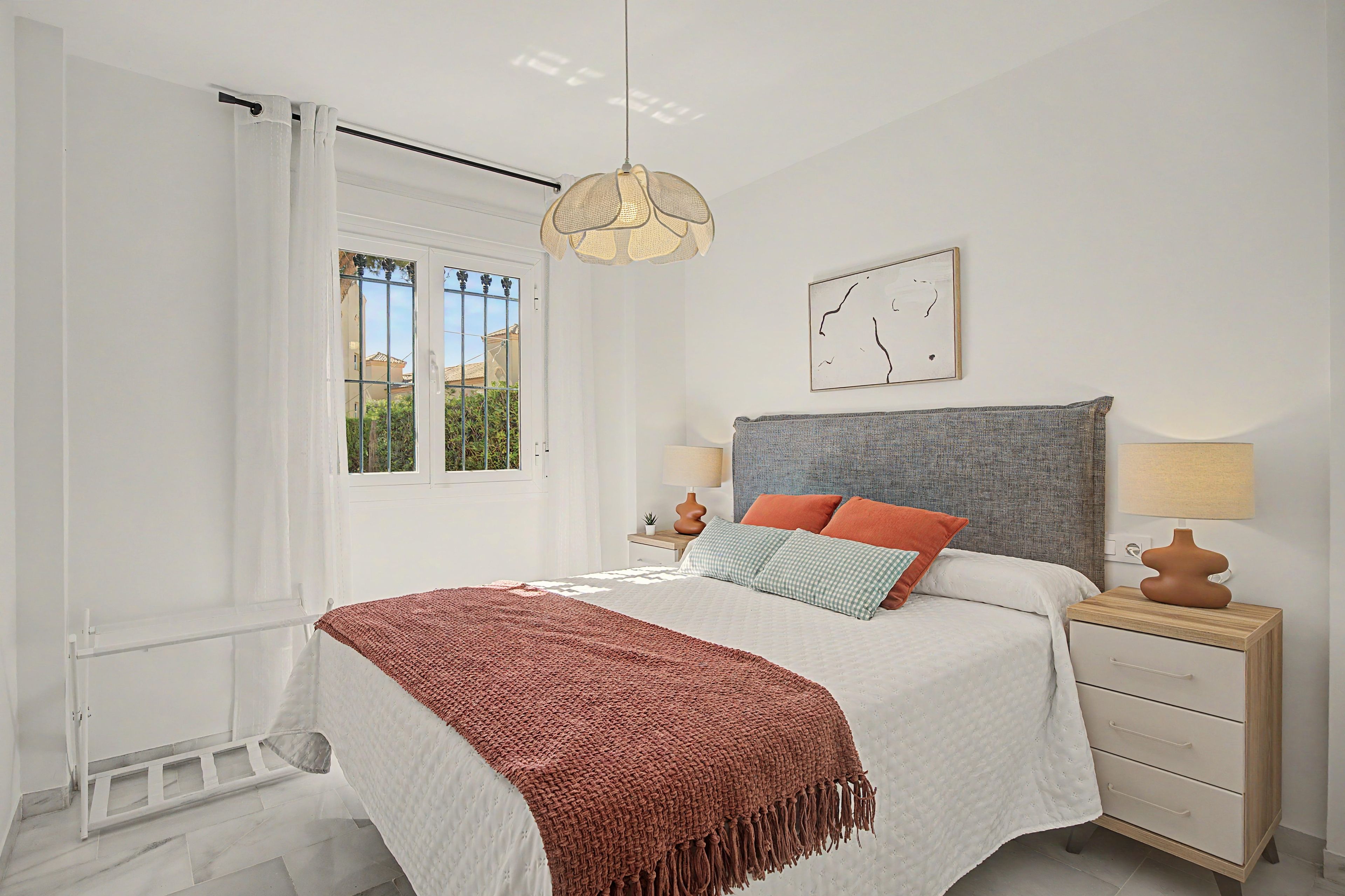 Un dormitorio con cama, mesitas de noche, lámpara colgante, ventana y una obra de arte en la pared.