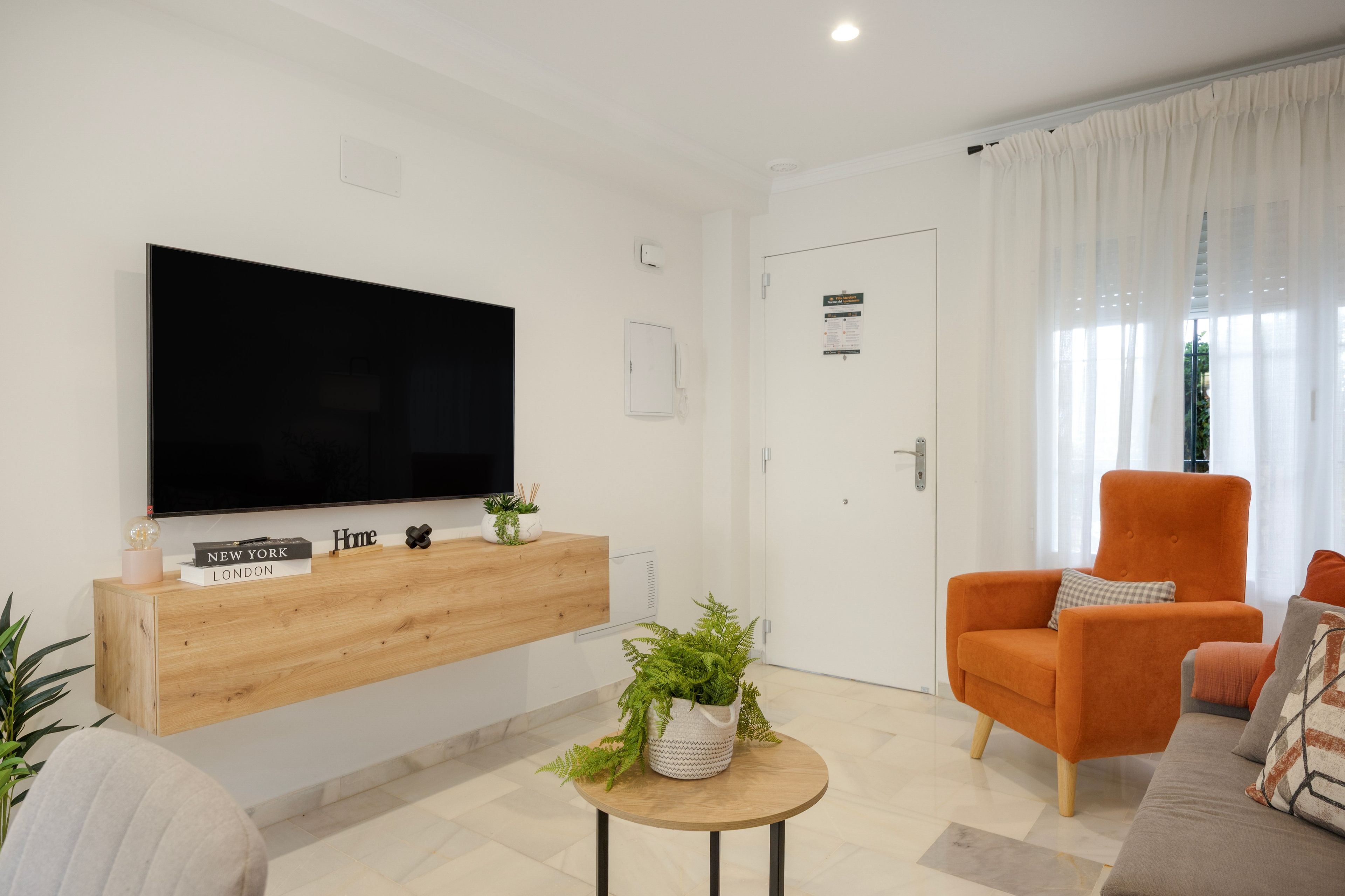 Ett modernt vardagsrum med en platt-TV, ett träbord, ett runt soffbord med en växt, en orange fåtölj och en grå soffa.