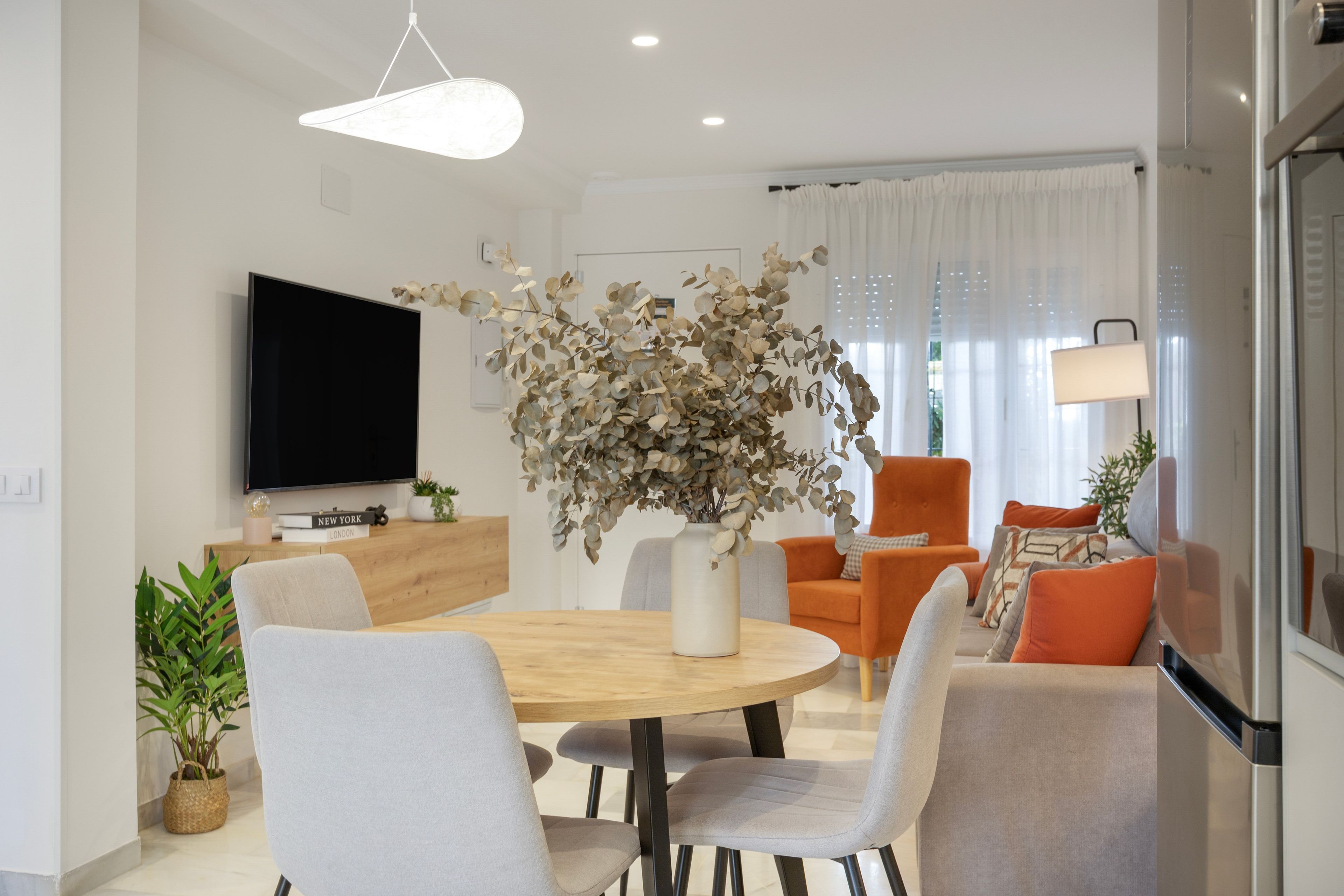A modern living room with a round wooden dining table, grey upholstered chairs, a flat-screen TV mounted on the wall, and a large vase with dried eucalyptus branches.