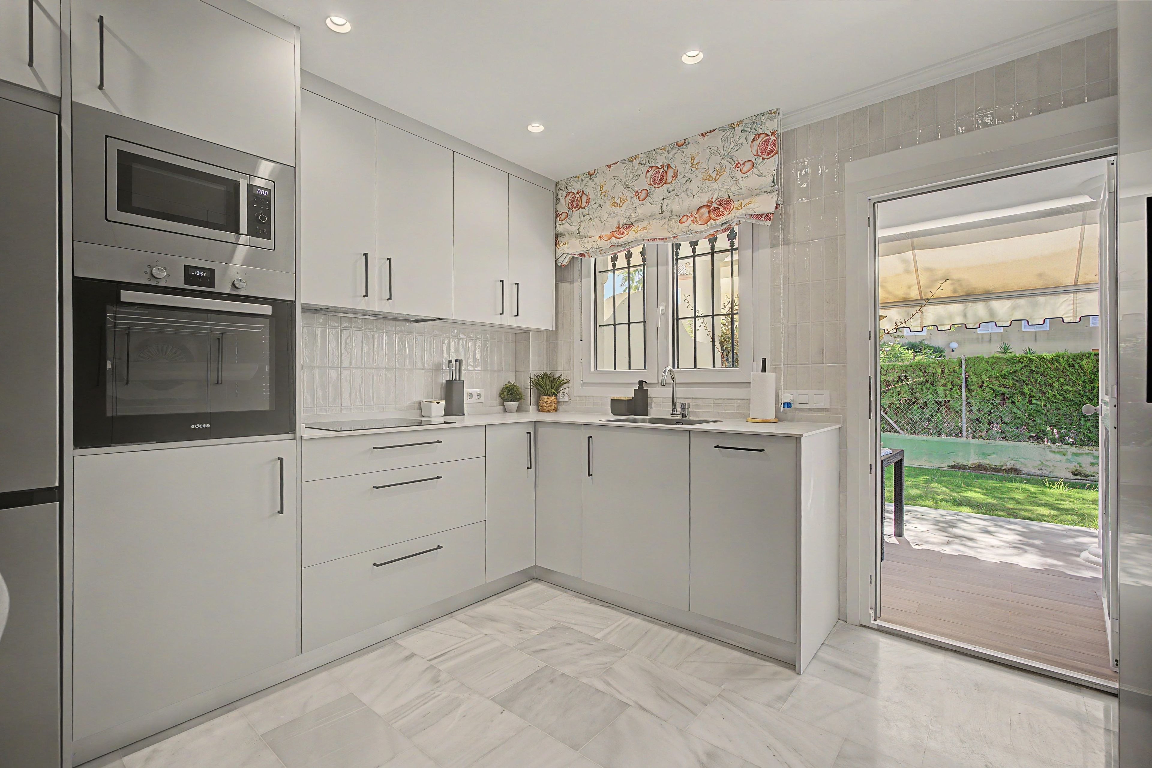 Une cuisine moderne avec des meubles blancs, un four encastré et une fenêtre agrémentée de rideaux décoratifs.