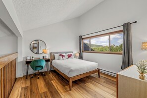 Ein Schlafzimmer mit Holzboden, einem Bett mit weißer Bettdecke, einem Schreibtisch mit Stuhl, einem Fenster mit Blick auf Grünflächen und einem an der Wand befestigten Spiegel.